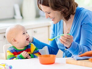 אמא ותינוק יושבים ליד שולחן בעוד האמא מחייכת לתינוקה ומחזיקה בידה כפית מלאה באוכל לארוחת בוקר