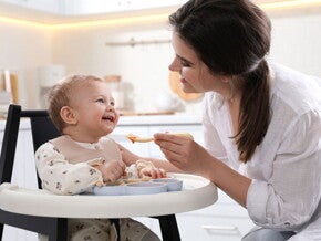 אמא מאכילה את תינוקה בכפית, כשהוא יושב בכסא האוכל שלו