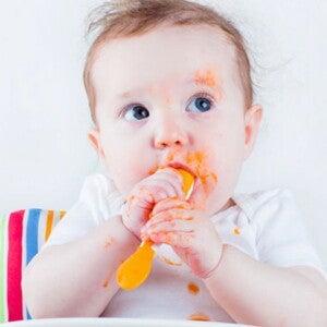 תינוק מחזיק כפית ומרוח במחית גזר בכל פניו