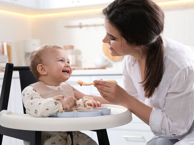 אמא מאכילה את תינוקה בכפית, כשהוא יושב בכסא האוכל שלו