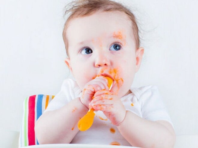 תינוק מחזיק כפית ומרוח במחית גזר בכל פניו