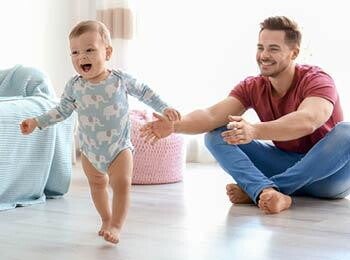 תינוקת בגיל שנה הולכת את צעדיה הראשונים ואביה שמח מאחוריה